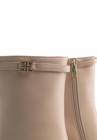 Stivaletti beige in pelle con un accento in metallo dorato e zip laterale. Texture liscia con dettagli cuciti lungo il bordo superiore.