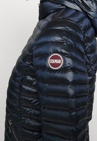 Giacca a vento in navy con design trapuntato, texture lucida, tasca laterale con zip e patch rotondo sul braccio superiore.