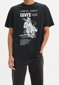 Czarna bawełniana koszulka z grafiką kowboja na koniu z lassem, białym napisem i postrzępionymi brzegami. Noszona z czarnymi dżinsami.