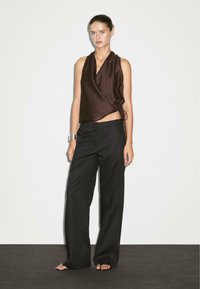 Le mannequin se tient devant un fond neutre, portant un haut enveloppant marron foncé et un pantalon noir ample, mettant en avant un style à la fois décontracté et élégant.