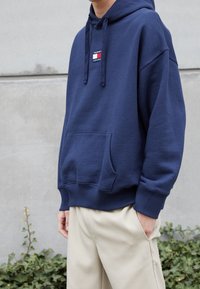 Felpa blu navy con tasca anteriore, cappuccio con cordino e patch con logo. Vestibilità comoda con polsini e orlo a coste, abbinata a pantaloni beige.