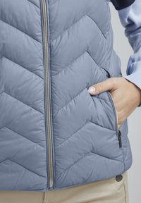 Vesta cu un model în zigzag, de culoare albastru deschis, cu fermoar frontal și buzunare laterale. Material moale cu o textură netedă, având accesorii din metal argintiu.