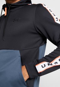 Sort og blå sports hættejakke med høj krave, lynlås og kontrastfarvede orange detaljer. Har brandede striber langs ærmerne.