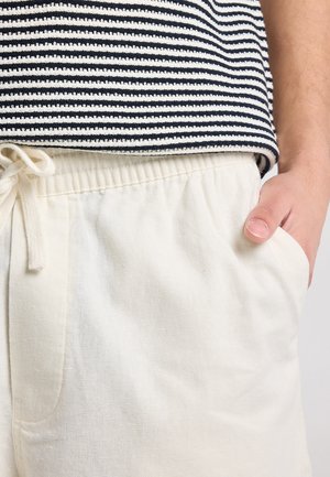 Personne portant un pantalon blanc à cordon avec la main dans la poche et une chemise rayée noir et blanc rentrée à l'intérieur.