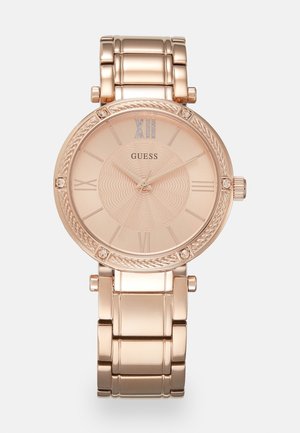 Montre bracelet en métal or rose avec chiffres romains, cadran texturé, accents en cristal et logo "GUESS" sur le cadran.