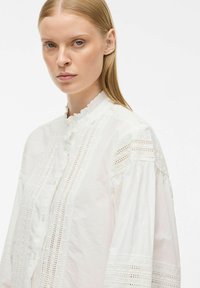 Witte katoenen shirt met een hoge kraag, voorzien van ingewikkelde kantinvoegen, geplooide mouwen en knoopsluiting aan de voorkant.