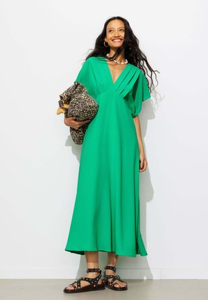 Femme en robe vert vif tenant une veste imprimée léopard, portant des sandales noires cloutées et souriant devant un fond blanc.