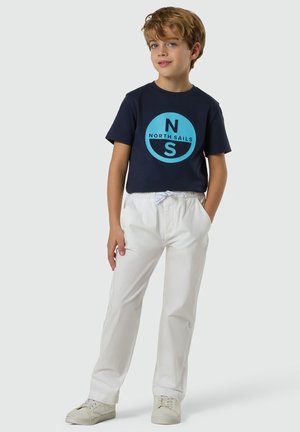 Junge steht mit Händen in den Taschen, trägt ein dunkelblaues North Sails T-Shirt, weiße Hose und weiße Turnschuhe vor einfarbigem Hintergrund.