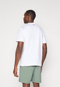 Λευκό κοντομάνικο μπλουζάκι με στρογγυλή λαιμόκοψη και μαύρο λογότυπο πίσω, συνδυασμένο με πράσινες βερμούδες. Λείος υφασμάτινος χαρακτήρας, casual εφαρμογή.