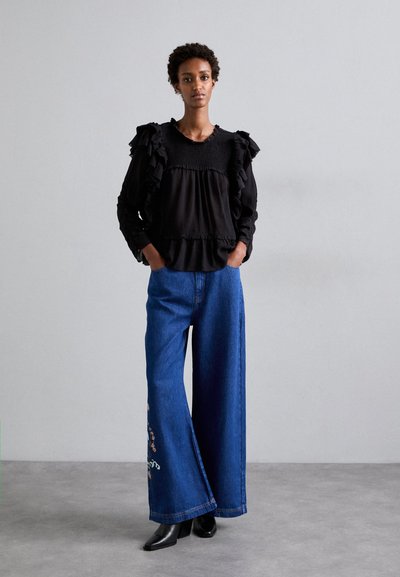 Black ruffled blouse with a smocked bodice, paired with wide-leg blue denim jeans featuring floral embroidery and black heeled boots.