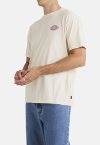 Camiseta de manga corta de color beige en tejido de algodón, que presenta un gráfico de un ojo rojo y azul en el lado izquierdo del pecho. Etiqueta en la costura lateral en la parte inferior izquierda.