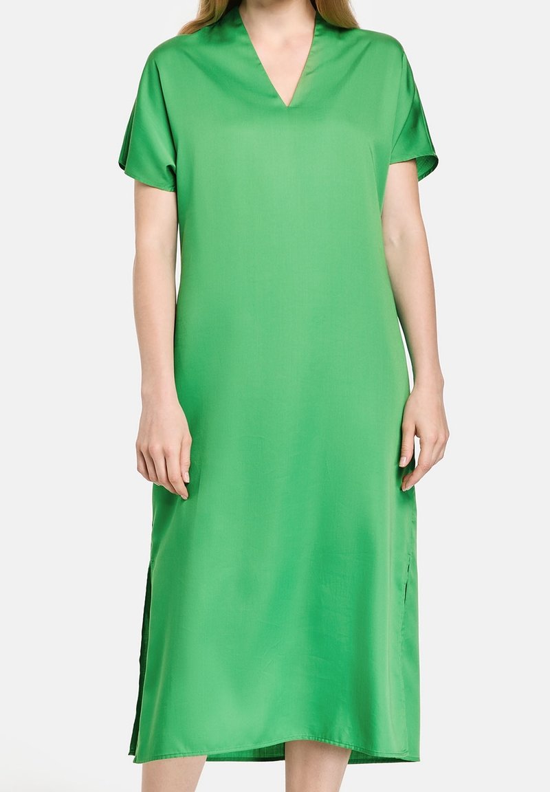 Femme portant une robe verte vif longueur genou avec manches courtes et un décolleté en V, debout devant un fond clair uni.