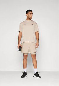 Camiseta deportiva beige con el logo «CNZ», combinada con pantalones cortos a juego. El modelo sostiene una botella de agua negra y lleva zapatillas negras.