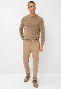 Bruin gestructureerd gebreid trui met een ribbelontwerp, gecombineerd met lichtbruine chino's en bruine suède schoenen. Model staat tegen een witte achtergrond.