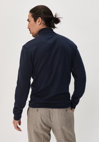 Polo de marină cu mânecă lungă, guler înalt și manșete cu nervuri, asortat cu pantaloni texturați gri deschis, prezentat din spate.
