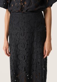 Jupe en dentelle noire ornée de motifs floraux complexes, avec une taille cintrée et une silhouette fluide. La texture est délicate et translucide.