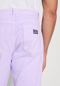 Fialové bavlněné kalhoty s přímým střihem, s dvěma zadními kapsami a malou černobílou logou na pravé kapse.