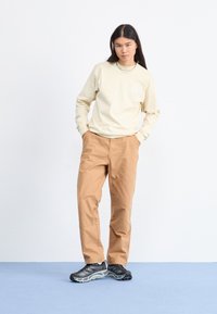 Chemise crème à manches longues avec un petit logo en triangle, associée à un pantalon cargo beige et des chaussures de sport noires avec des accents gris. Coupe simple et décontractée.
