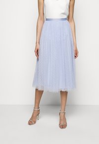 Kvinna som bär en ljusblå midi-tullkjol med vita prickar, en vit ärmlös topp och silverfärgade högklackade sandaler med ankelremmar.