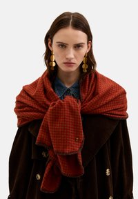 Rød ternet tørklæde lavet af blødt stof, lagt over en mørkebrun kordjakke med et guldemblem og frynser i kanterne.