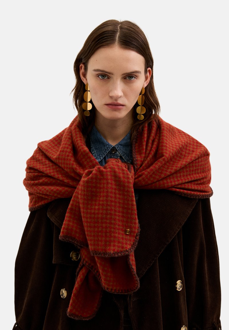 Rød ternet tørklæde lavet af blødt stof, lagt over en mørkebrun kordjakke med et guldemblem og frynser i kanterne.