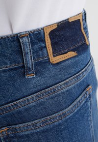 Mörkblå denimjeans har en brun läderlapp, synlig sömnad och en standard femfickdesign. Tyget har en slät textur.