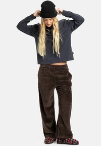 Donkergrijze cropped sweatshirt met lange mouwen, gecombineerd met bruine corduroy broek en rood geruite slippers. Eenvoudige, textuur rijke materialen zijn aanwezig.