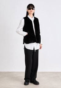 Schwarzer Kunstpelz-Westen mit zwei Seitentaschen, getragen über einem weißen Hemd, kombiniert mit schwarzen Hosen und klobigen schwarzen Schuhen. Schlicher Hintergrund.