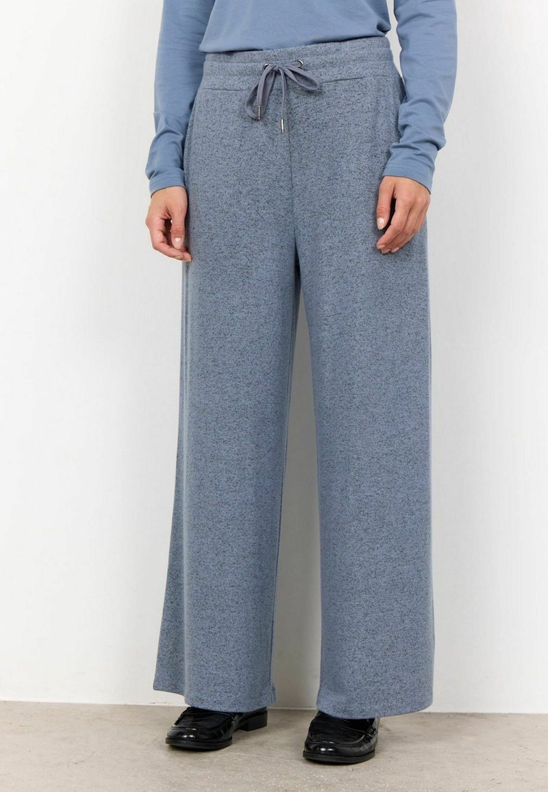 Personne portant un pantalon large gris-bleu avec un cordon de serrage à la taille, un haut à manches longues bleu clair et des chaussures noires, se tenant à l'intérieur.