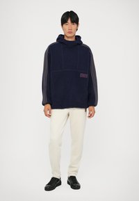 Camisola de polar azul marinho com capuz, textura ribajada e riscas laterais. Apresenta um bolso frontal e um pequeno emblema perto do bolso.