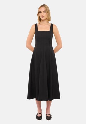 Femme aux cheveux blonds courts portant une robe midi noire sans manches avec un décolleté carré et des chaussures Mary Jane noires, debout les mains dans le dos.