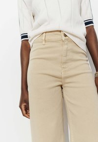 Pantalon beige taille haute en denim avec une texture lisse et un design classique à cinq poches. Comprend une fermeture éclair à l'avant et un bouton.