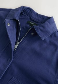 Tamsiai mėlyna marškinėliai-striukė su tekstūruota audinio faktūra, su užtrauktuku, spaudėmis ir apykakle. Etiketėje parašyta „NEXT“.