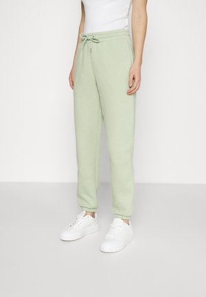 Person trägt hellgrüne Sweatpants mit elastischen Bündchen, weiße Plateau-Sneaker und ein weißes ärmelloses Oberteil vor einem einfarbigen Hintergrund.