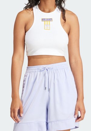 Femme portant un haut court sans manches blanc Adidas et un short à cordon de serrage violet clair, debout les bras détendus le long du corps.
