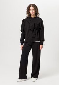 Schwarzer oversized Sweatshirt und passende weit geschnittene Hose aus weichem Stoff, mit einer oberen Lage, die lässig über eine Schulter drapiert ist. Weiße Schuhe.