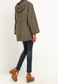 Olivově zelená bunda s kapucí, spadlými rameny a stahovacími šňůrkami v pase. Kombinováno s tmavými džíny a hnědými platformovými botami. Pohled ze zad.