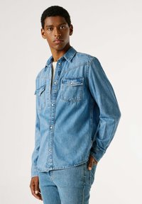 Světlomodrá denimová košile se dvěma náprsními kapsami, zapínáním na knoflíky a dlouhými rukávy. Látka má měkkou texturu a mírně opotřebovaný vzhled.