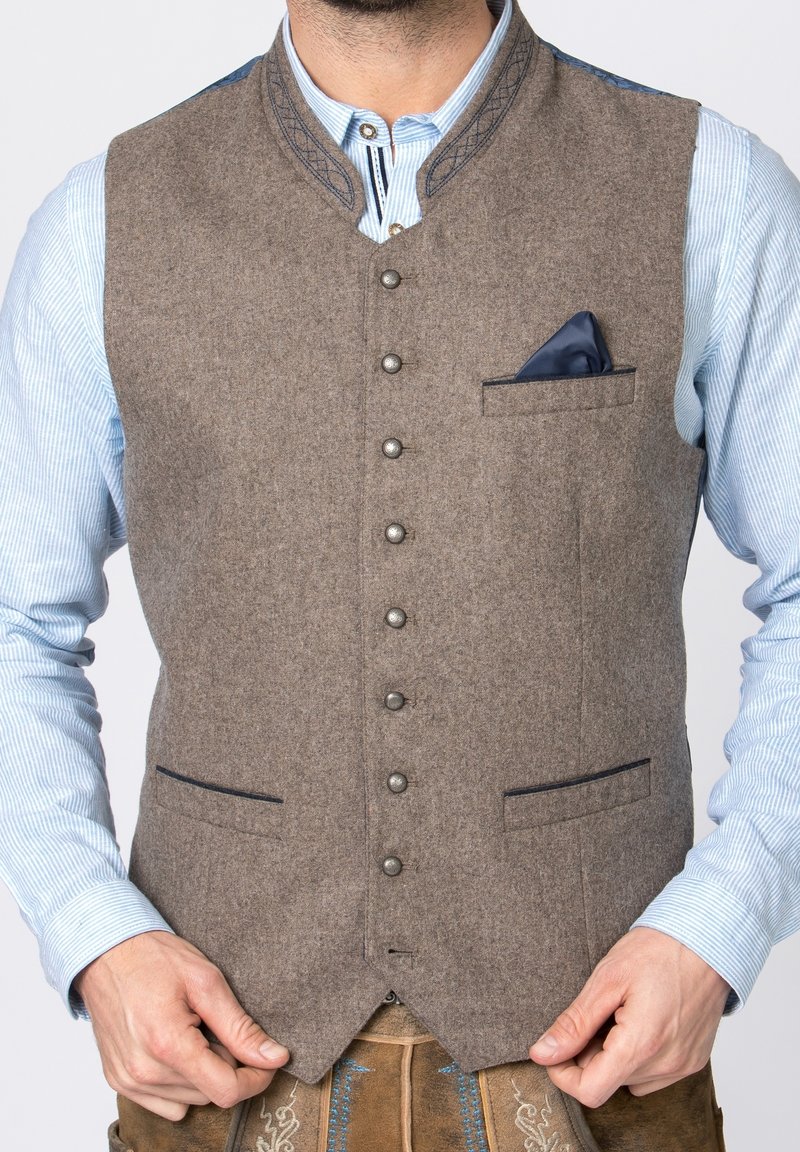 Homme portant un gilet en laine marron boutonné avec un col brodé, par-dessus une chemise bleu clair à rayures et un pantalon en cuir traditionnel.