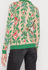 Pull en tricot beige avec des motifs floraux en rouge, bleu et vert. Présente un col et un ourlet côtelés verts. Le design s'étend sur le dos.