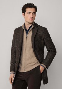 Abrigo de lana a cuadros en marrón oscuro con acentos en rojo y verde sobre un suéter beige con cremallera. La modelo posa con una mano en el bolsillo, con pantalones oscuros visibles.