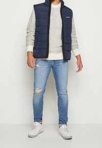 Marineblaues Pufferwesten über einem hellgrauen Pullover mit gerippten Bündchen, kombiniert mit lässigen, leicht ausgefransten hellblauen Jeans und weißen Sneakers, die einen entspannten Look präsentieren.