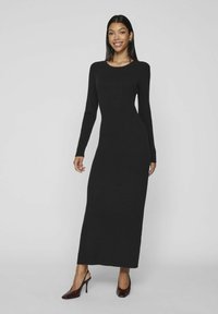 Musthave mustribaldatud pikkade varrukatega kleit ümmarguse kaelusega, keha lähedane lõige ja pahkluu pikkus, koos tumedate pruunide kontsakingadega.