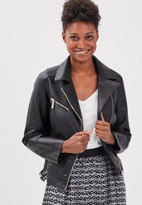 Giubbotto da motociclista in pelle sintetica nera con accessori in argento, tasche con zip e design con colletto aperto, indossato sopra una maglia bianca e una gonna a fantasia.