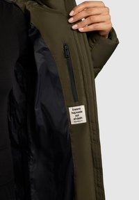 Olivovo zelená prešívaná bunda s čiernou nylonovou podšívkou, s bočnou zipsovú kapsou a látkovou nálepkou, na ktorej je nápis "Teplé objatie vyrobené z látky."