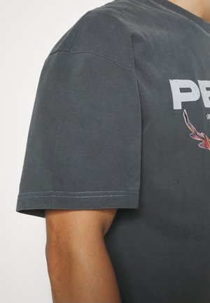 Osoba ubrana w ciemnoszary t-shirt z krótkim rękawem, z częściowym białym napisem i małym kolorowym wzorem na klatce piersiowej.