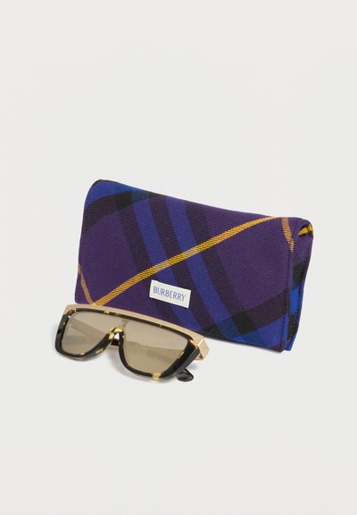 Γυαλιά ηλίου με σκελετό από χελώνα και μπεζ φακούς, τοποθετημένα μπροστά σε μια θήκη Burberry με μοτίβο καρό σε μωβ, μπλε και κίτρινο.