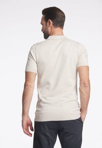 Kortärmad, beige stickad pikétröja med låg krage och ribbade ärmslut. Har en mjuk yta och en figursydd silhuett.