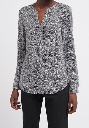 Blouse à manches longues tachetée noir et blanc avec un col en V et des boutons, portée avec un pantalon noir sur une personne debout.