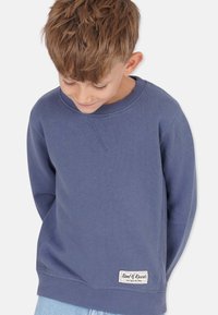 Sininen puuvillapaita, jossa on pyöreä kaula-aukko, resorit hihoissa ja helmassa. Vasemmassa alareunassa on pieni brändilogo.